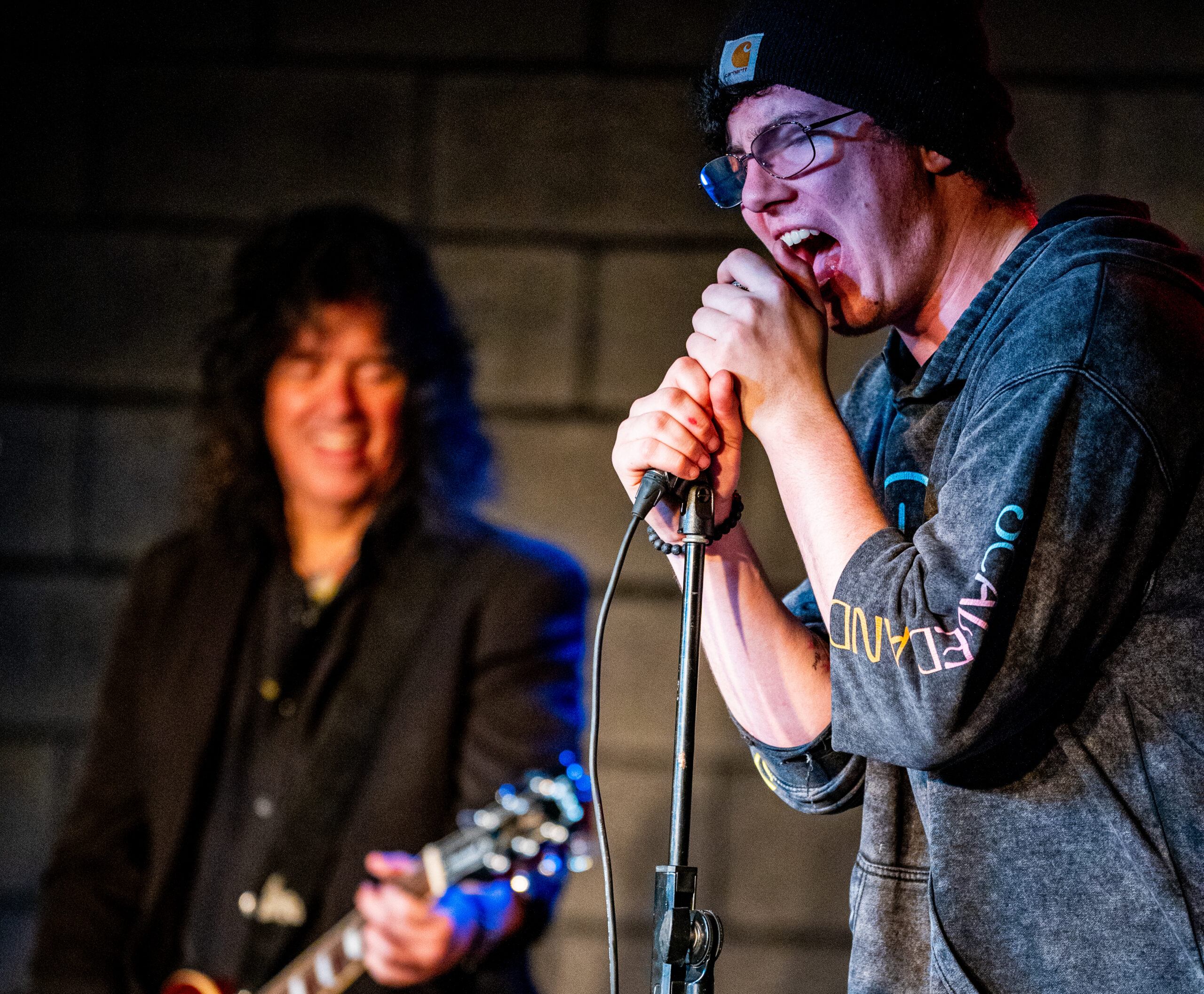 A high-energy moment during a live music performance. The singer is passionately singing into a microphone, wearing a hoodie and beanie, while gripping the mic stand. Behind him, a guitarist smiles while playing an electric guitar. The background is dimly lit and the focus on the musicians.