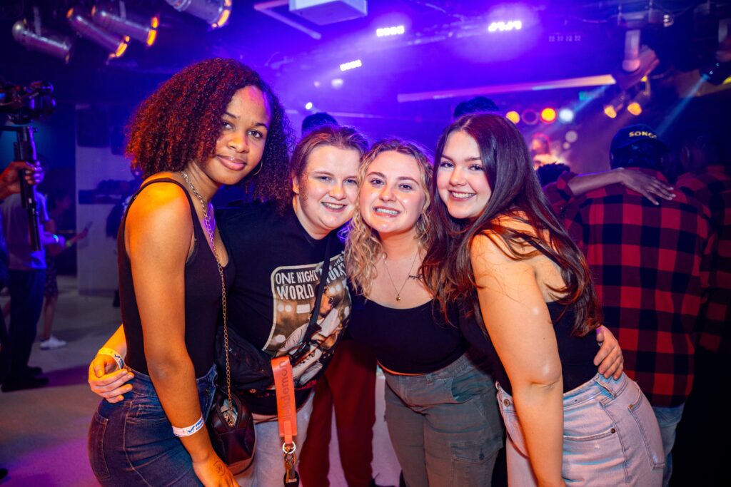 Four female students stand huddled together smiling at the camera. The background is dimly lit with purple lighting. Other students are visible in the background, showing a busy pub scene.