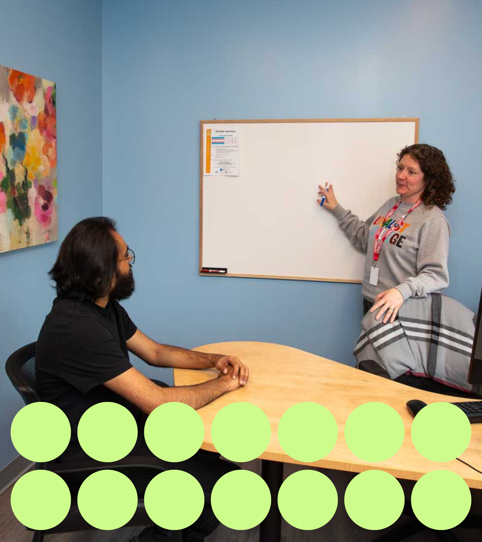 A student sits across from a counsellor in a counselling room. The counsellor, wearing a Loyalist College sweatshirt, gestures at a whiteboard on the wall while engaging with the student. A graphic with green dots is at the bottom of the image.