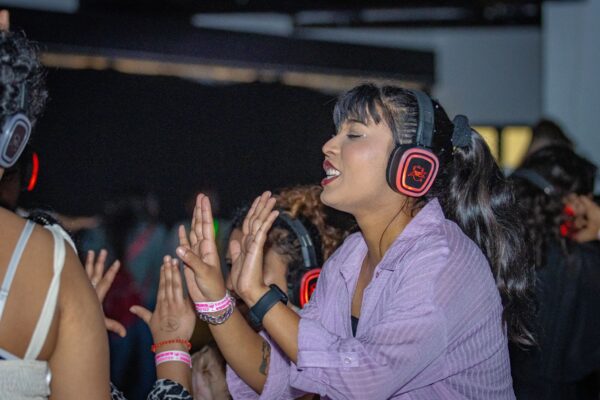 A person enjoying a silent disco, wearing headphones and dancing with enthusiasm. The vibrant lighting and purple shirt add to the lively atmosphere, as they raise their palms, fully immersed in the music.