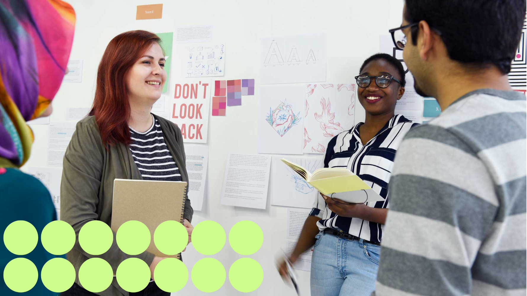 Group of Students Standing in a circle, holding notebooks and talking to one another in a classroom. The classroom has graphics and sketches on the wall