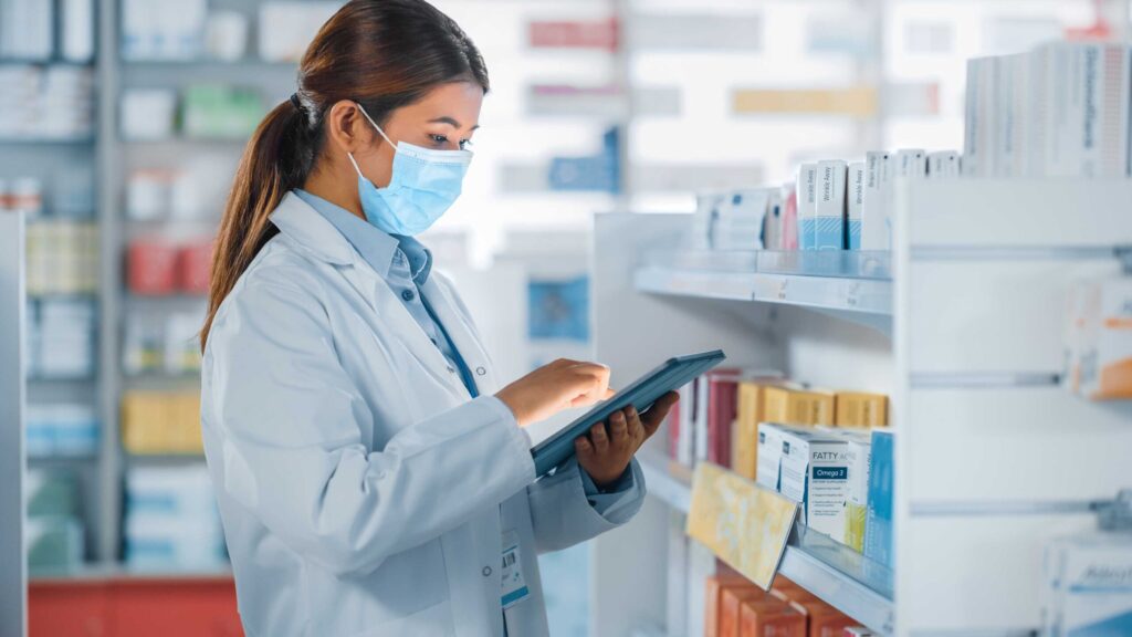 A pharmacy technician wearing a white lab coat and a face mask uses a tablet while managing pharmacy inventory. Shelves filled with pharmaceutical products line the background.