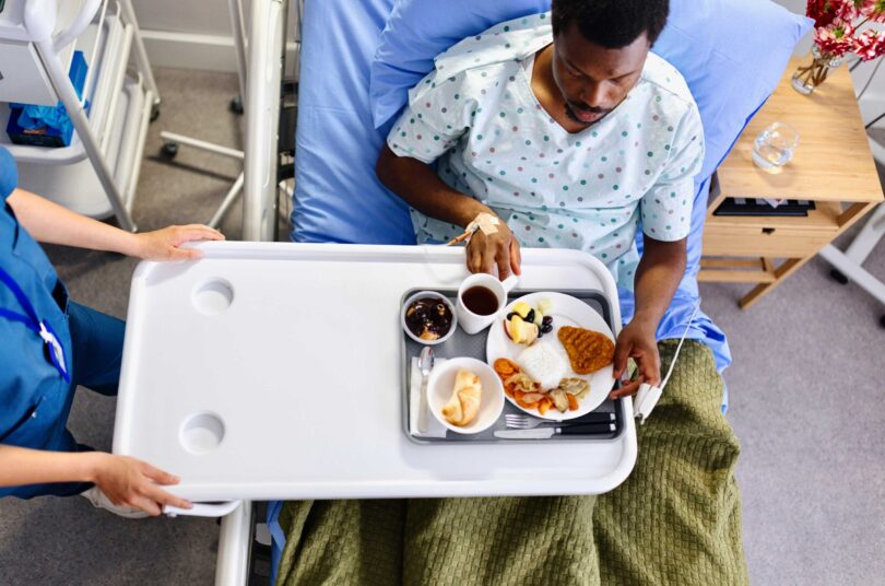 A food service worker brings a meal served on a tray to a patient in a health care setting.