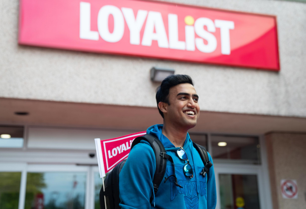 Male Loyalist College student standing out in front of a building. He's smiling and starring off camera, wearing a backpack with a Loyalist pennant on the side.