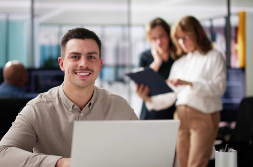 Smiling man wearing professional attire works on a laptop in an office environment.