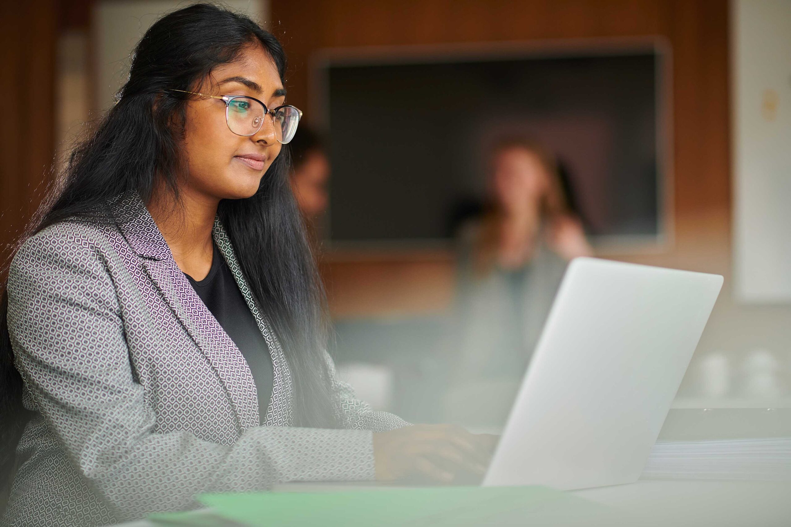 Woman wearing glasses and professional attire works on a laptop in an office environment.