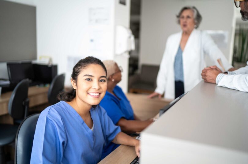 Smiling woman wearing medical uniform sits at a front reception desk on a computer in a medical office setting. In the background are other members of staff, including doctors.