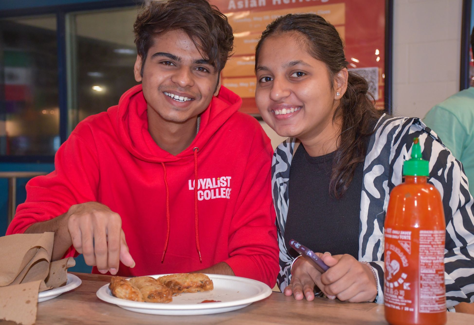 Two smiling students, a young man and a young woman, at a spring roll making workshop in a college cafeteria. The young man, wearing a red hoodie with 'LOYALIST COLLEGE' printed on it, points to a plate of freshly made spring rolls in front of him. The young woman, wearing a zebra-print cardigan over a black top, holds a phone in one hand, ready to capture the moment. A bottle of hot sauce is visible on the table, adding a spicy touch to the setup.