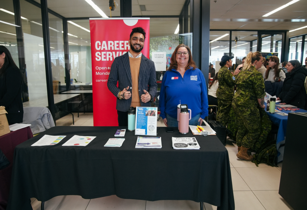 Two members of the Career Services staff standing behind a table at an information centre. On the table there are several printed materials. In the background there are other booths with both staff and students around talking.