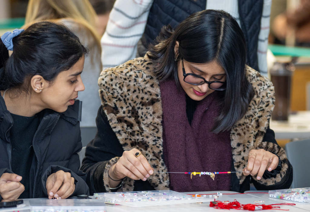 Two students sit at a table covered in beads and string, focused on creating handmade bracelets. One student holds up a beading needle strung with colourful beads, while the other looks on and offers input. The atmosphere is relaxed and creative, with other participants visible in the background.