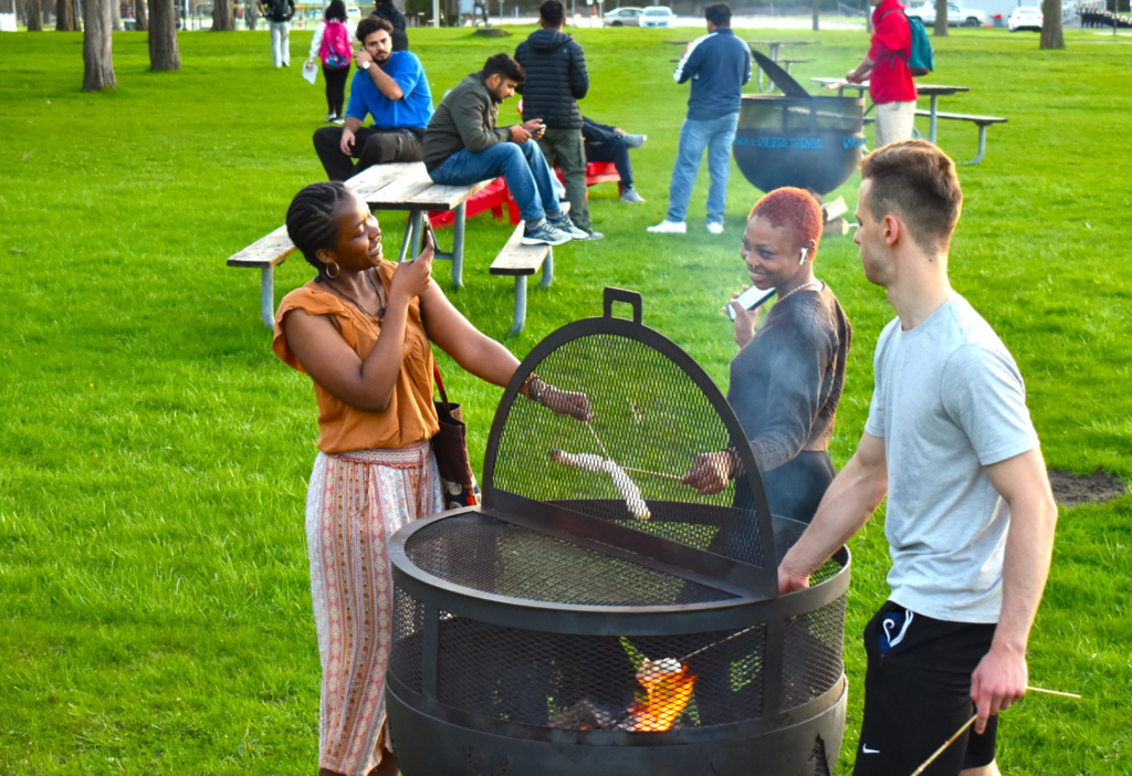 Students gather outdoors in a green park for an evening campfire. Three students stand around a fire pit roasting marshmallows, chatting and laughing, while others relax on picnic tables in the background. The scene captures a sense of community and fun during a casual campus event.