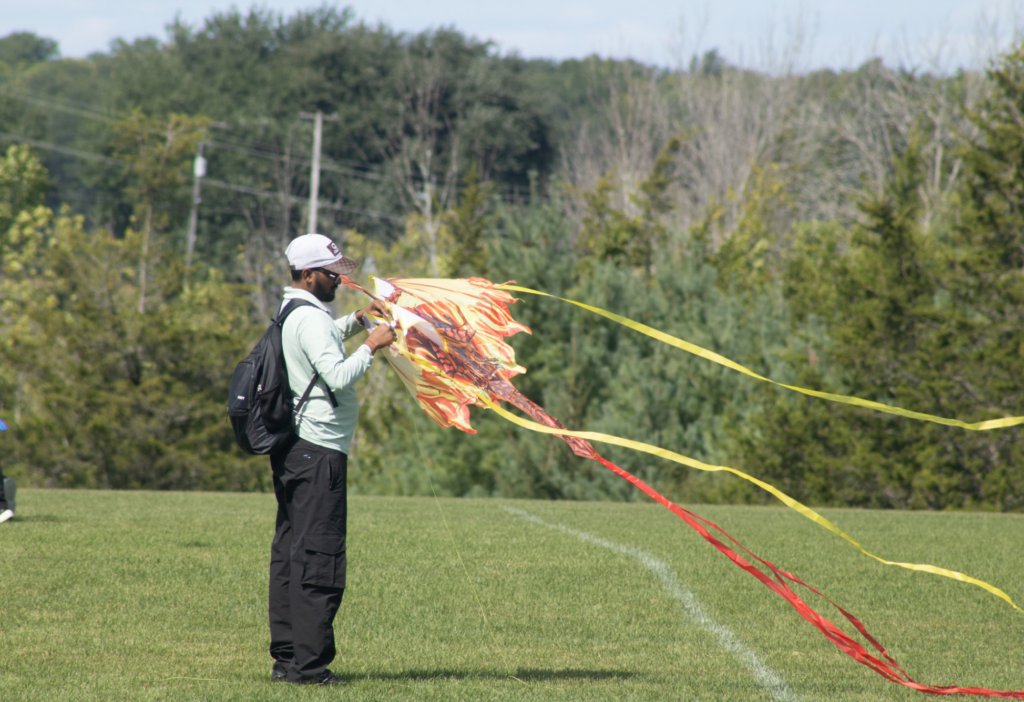 A person stands on a grassy field holding a vibrant, flame-patterned kite with long red and yellow streamers blowing in the wind. They are wearing a white cap, sunglasses, a light green shirt, black cargo pants, and a black backpack, with trees and power lines in the background.