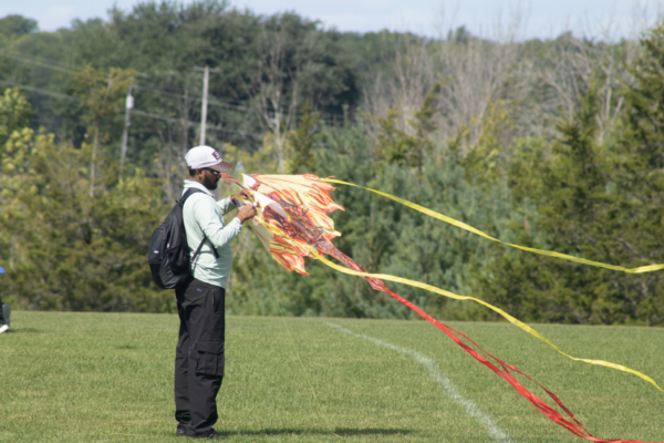 A person stands on a grassy field holding a vibrant, flame-patterned kite with long red and yellow streamers blowing in the wind. They are wearing a white cap, sunglasses, a light green shirt, black cargo pants, and a black backpack, with trees and power lines in the background.