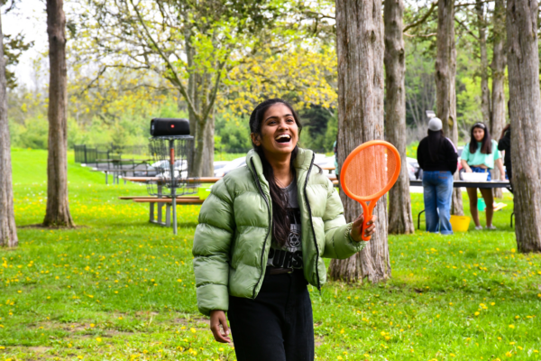 A student wearing a green puffer jacket smiles brightly while holding a plastic paddle during an outdoor game surrounded by tall trees. In the background, other students gather near a table with activity supplies, and disc golf baskets are visible among the picnic benches, creating a playful and engaging atmosphere.