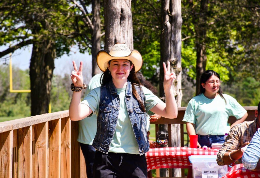 A student wearing a cowboy hat and denim vest poses with both hands up in peace signs at a sunny outdoor event. Behind her, other students engage in activities at picnic tables covered in red checkered tablecloths, with signs like “DIY Tote Bags” adding to the playful, community vibe.