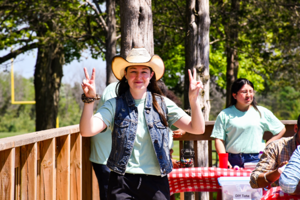 A student wearing a cowboy hat and denim vest poses with both hands up in peace signs at a sunny outdoor event. Behind her, other students engage in activities at picnic tables covered in red checkered tablecloths, with signs like “DIY Tote Bags” adding to the playful, community vibe.