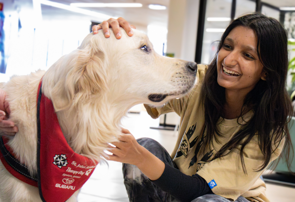 A smiling student gently pets a golden retriever wearing a red bandana that reads “St. John Ambulance Therapy Dog.” The interaction takes place indoors, with the student visibly joyful, highlighting the calming and uplifting effect of the visit. The setting suggests a wellness or mental health support event on campus.