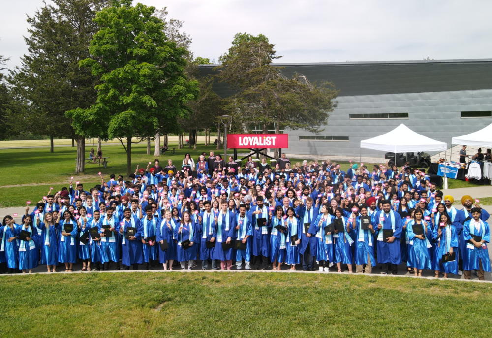 A large group of college graduates dressed in blue gowns and sashes gather outdoors for a group photo in front of a red "LOYALIST" sign. Many are holding diplomas and waving small Loyalist College flags. Behind them, trees, a modern building, and white event tents are visible under a partly cloudy sky.