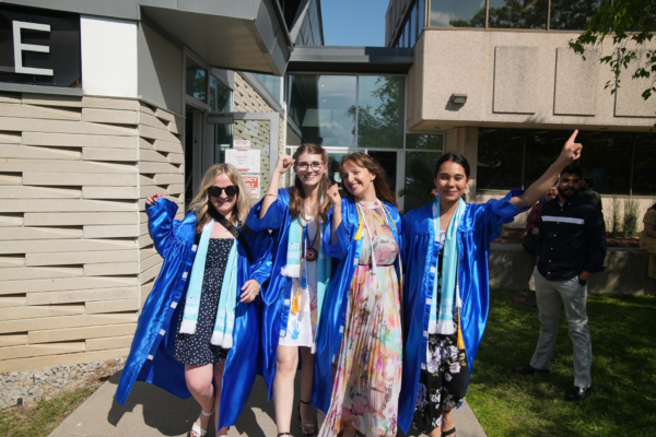 Four graduates wearing bright blue gowns and Loyalist Alumni scarves celebrate outside a building on a sunny day. They are smiling, holding hands, and making celebratory gestures. One person in the group is wearing a floral dress under her gown, while others wear dresses in various patterns. A few people stand in the background near the building.