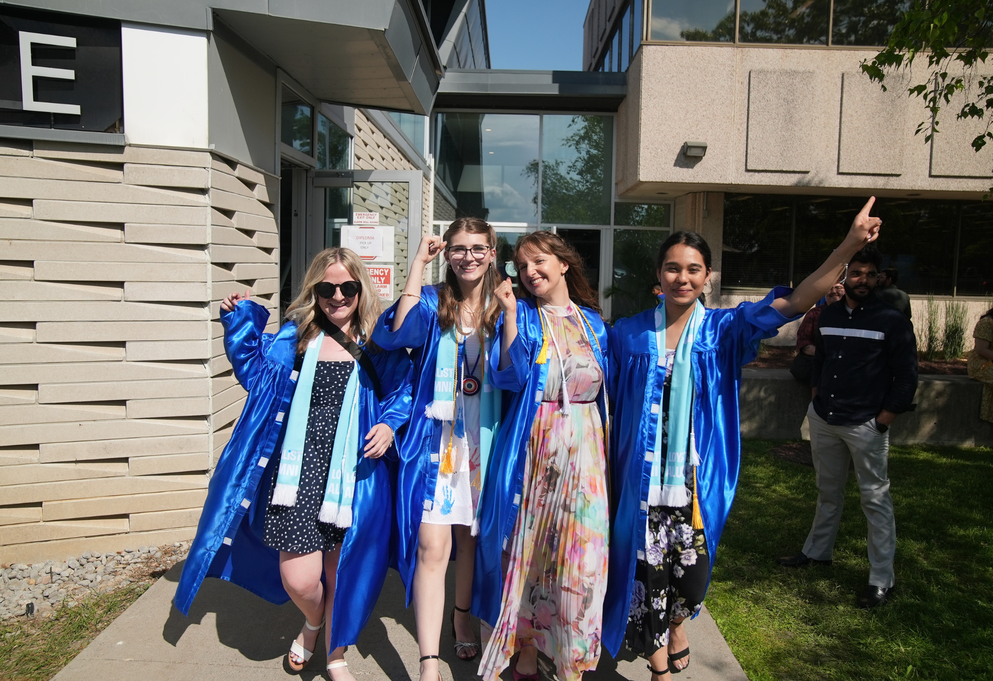 Four graduates wearing bright blue gowns and Loyalist Alumni scarves celebrate outside a building on a sunny day. They are smiling, holding hands, and making celebratory gestures. One person in the group is wearing a floral dress under her gown, while others wear dresses in various patterns. A few people stand in the background near the building.