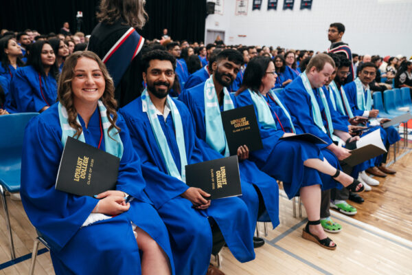 Group of Loyalist graduates wearing blue gowns and teal alumni scarves seated in a gymnasium. The graduates are holding black folders that say "Loyalist College Class of 2025".