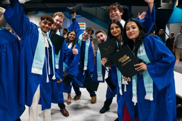 A group of Loyalist grads, all wearing blue gowns and teal alumni scarves. Most of them are holding black folders that say "Loyalist College Class of 2025" in gold lettering. They're all smiling and posing for the camera, some with their hands in the air.