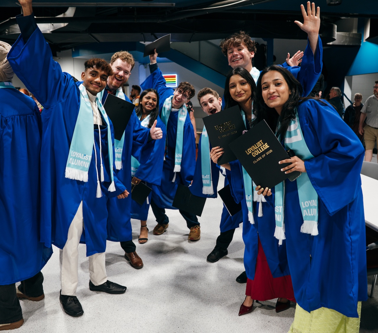 A group of Loyalist grads, all wearing blue gowns and teal alumni scarves. Most of them are holding black folders that say "Loyalist College Class of 2025" in gold lettering. They're all smiling and posing for the camera, some with their hands in the air.