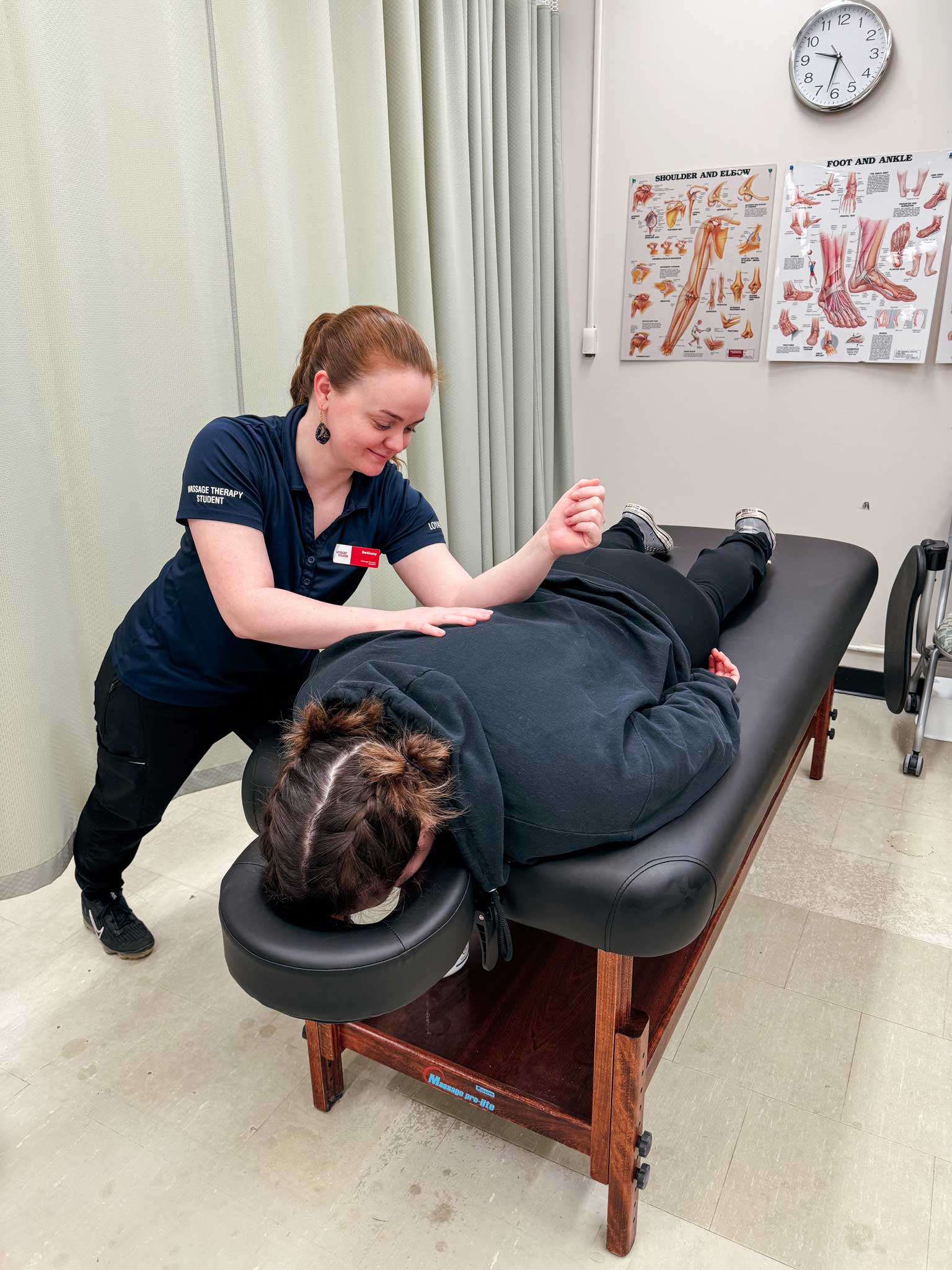 Female massage therapy student working on a patient face down on the massage bed in front of her. The student is bent over the patient, applying pressure on her back with her elbow. In the background are two posters of the muscular system on the wall, a clock, and a curtain blocking off the rest of the room.
