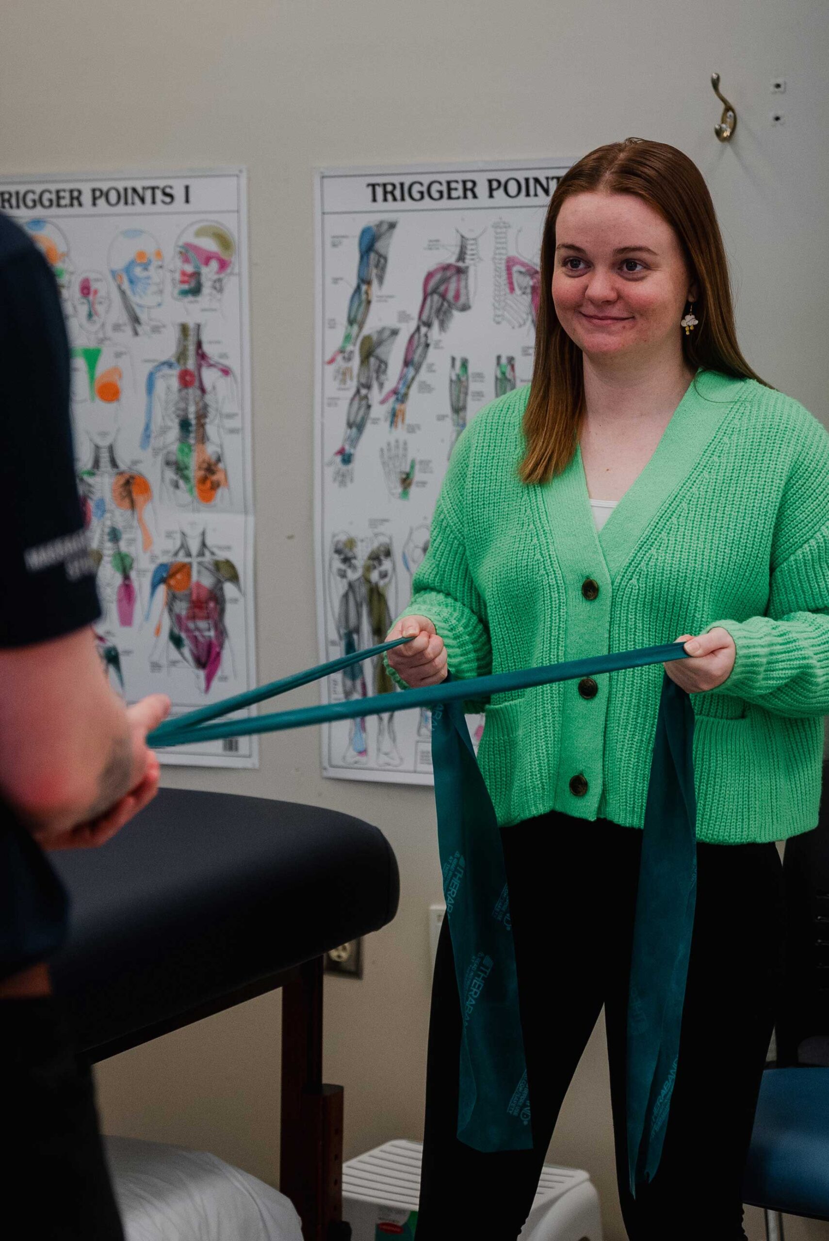 A girl in a bright green cardigan is pulling on elastic tension bands. She's smiling and looking at a figure standing just out of frame. In the background there are posters on the wall detailing trigger points on the body.