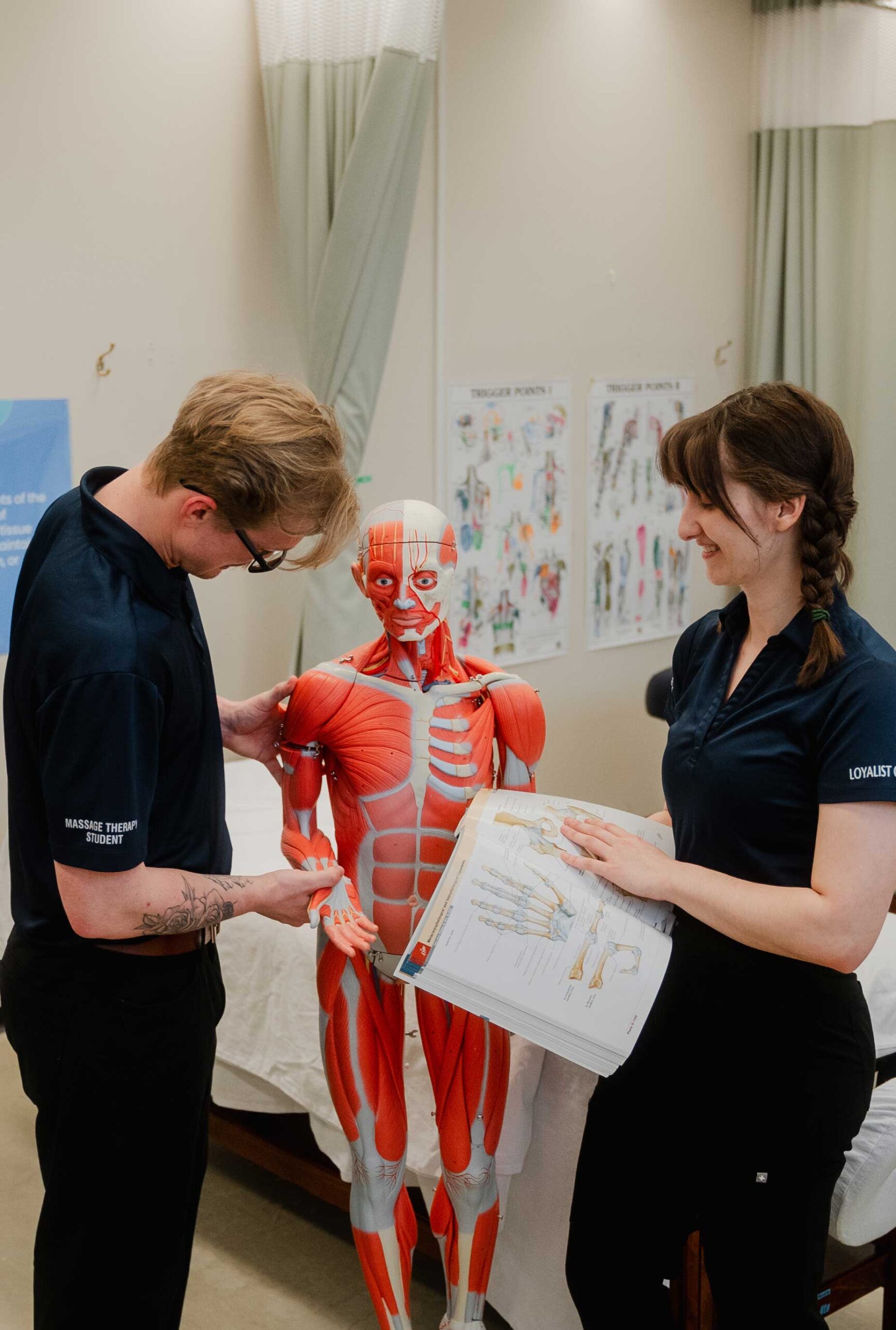 Two Loyalist Massage Therapy students are smiling in the Unwind Spa. The man is looking at a muscular skeleton model, while the woman is reading what appears to be a textbook on the skeletal system.