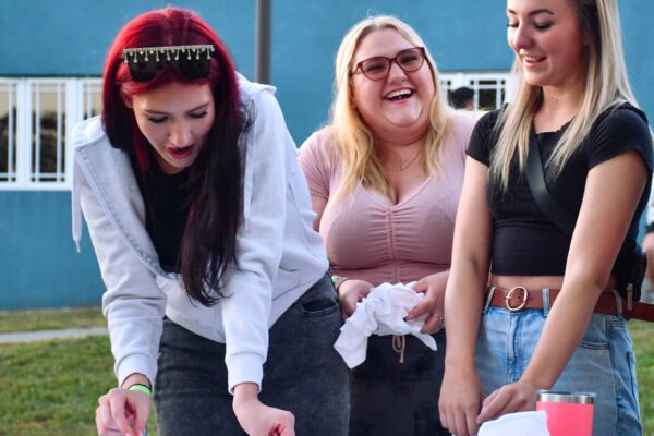 Three girls smiling and laughing outside. They're standing at a table while one girl is pinching a white t-shirt for tie-dying. There's another white shirt already bunched up on the table, and one girl is also holding a bundle.