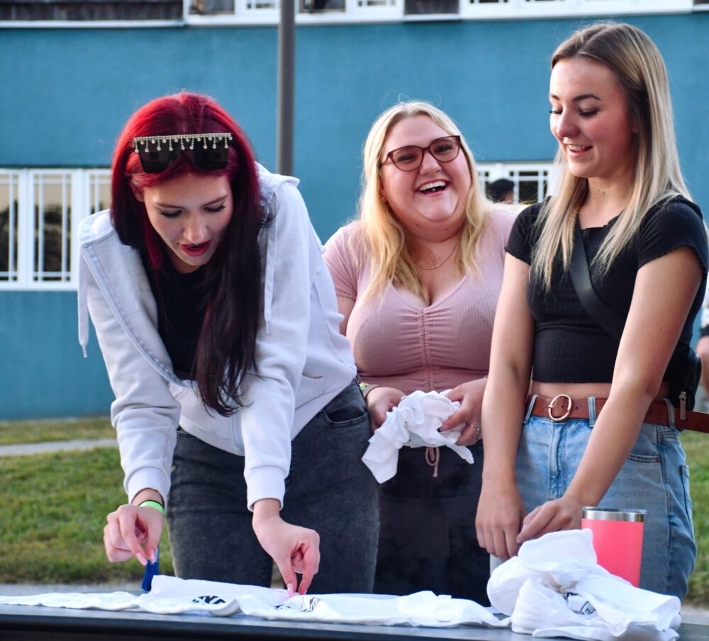 Three girls smiling and laughing outside. They're standing at a table while one girl is pinching a white t-shirt for tie-dying. There's another white shirt already bunched up on the table, and one girl is also holding a bundle.