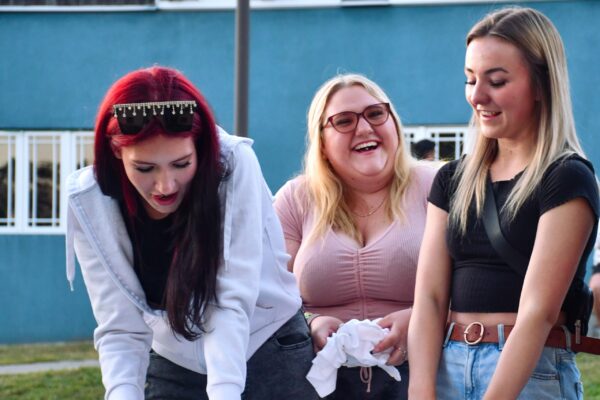Three girls smiling and laughing outside. They're standing at a table while one girl is pinching a white t-shirt for tie-dying. There's another white shirt already bunched up on the table, and one girl is also holding a bundle.