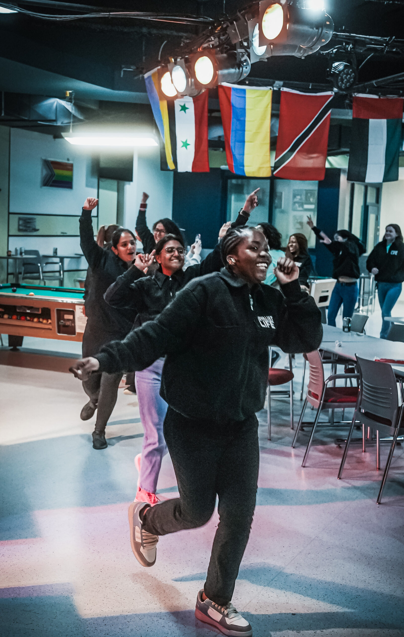 Students wearing black sweaters are dancing in a line in the Shark Tank Pub. There are flags for other countries hanging from the ceiling, pool tables and long tables with chairs in the background. The atmosphere is happy and fun.