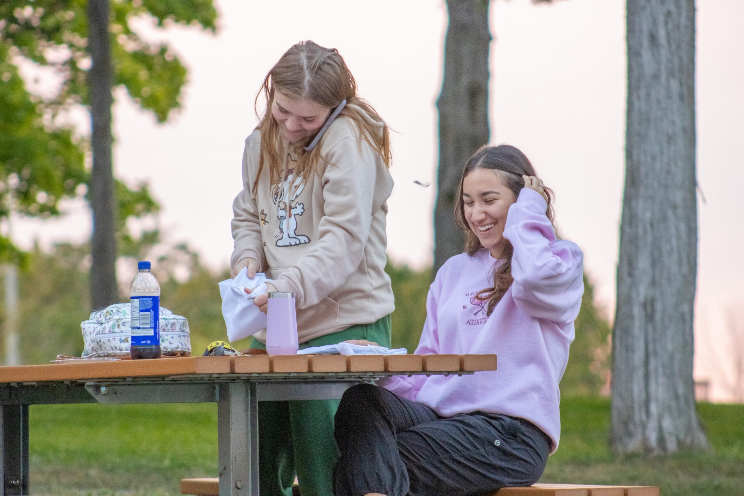 Two girls smiling and laughing at a picnic table. There's food and drinks in front of them, and one of the girls is standing with her phone to her ear.