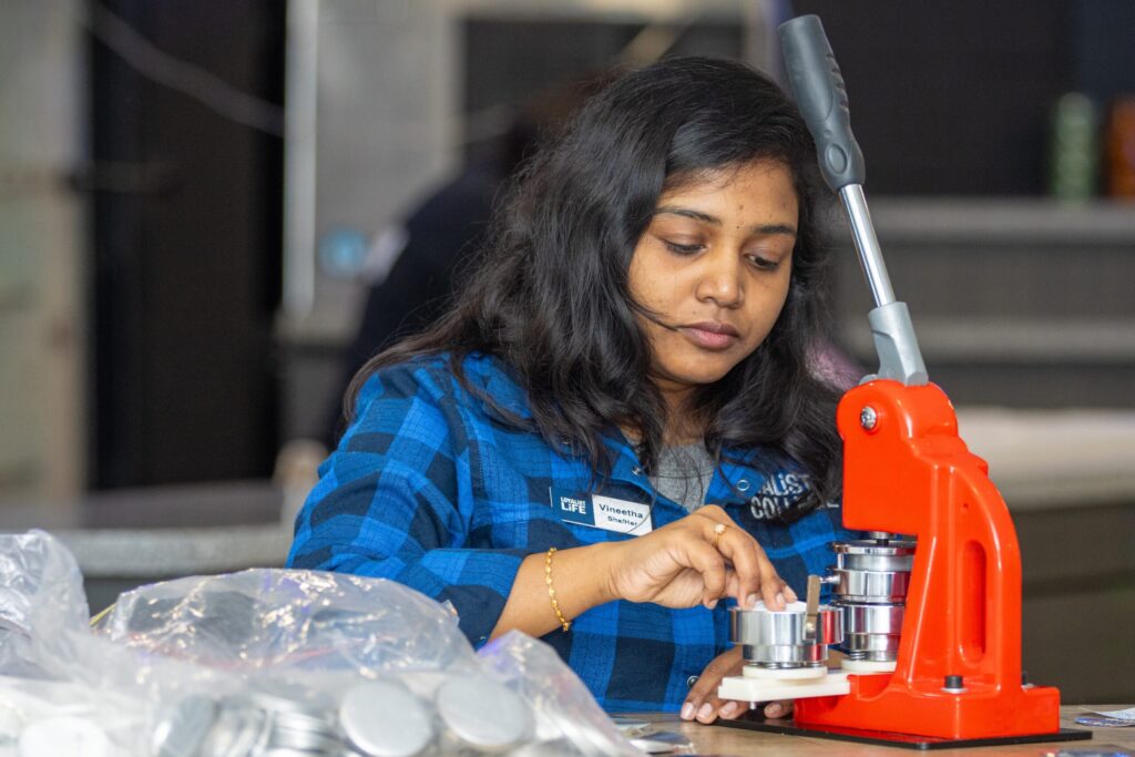 A female student wearing a blue plaid shirt and a Loyalist Life name tag is using a button maker. She's placing items into the machine, with a bag of more supplies to her left.