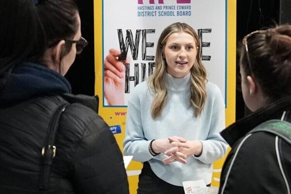 A representative speaks with attendees at a career fair booth with informational banners in the background.