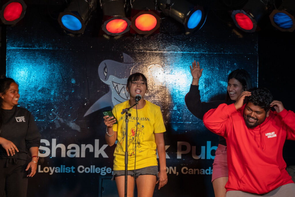 A group of 4 students on the stage in the Shark Tank Pub. One girl is standing at the mic and appears to be singing, while the other three appear to be dancing around her. Everyone is smiling.