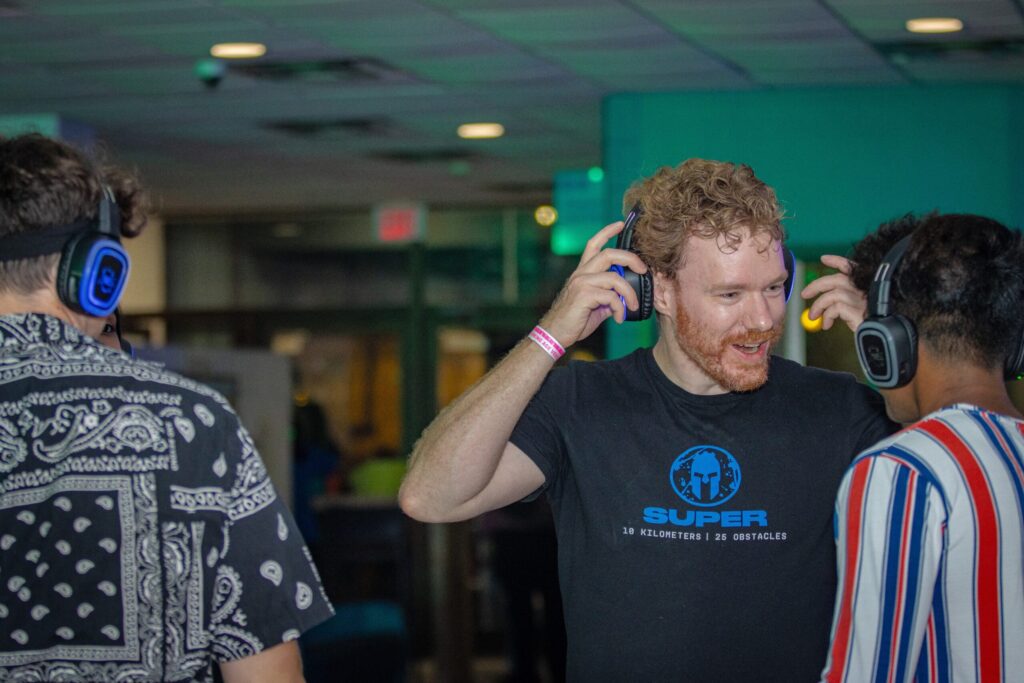 Male students at a pub night wearing silent disco headphones. One man is facing the camera and is smiling while lifting one side of the headphones to hear another man who appears to be speaking to him.