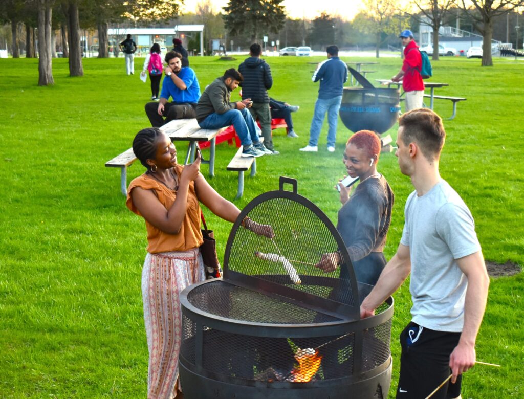 A group of students outside around firepits at Loyalist college. They're roasting marshmellows, taking pictures, talking and laughing. It's bright outside and the atmosphere is happy and fun.