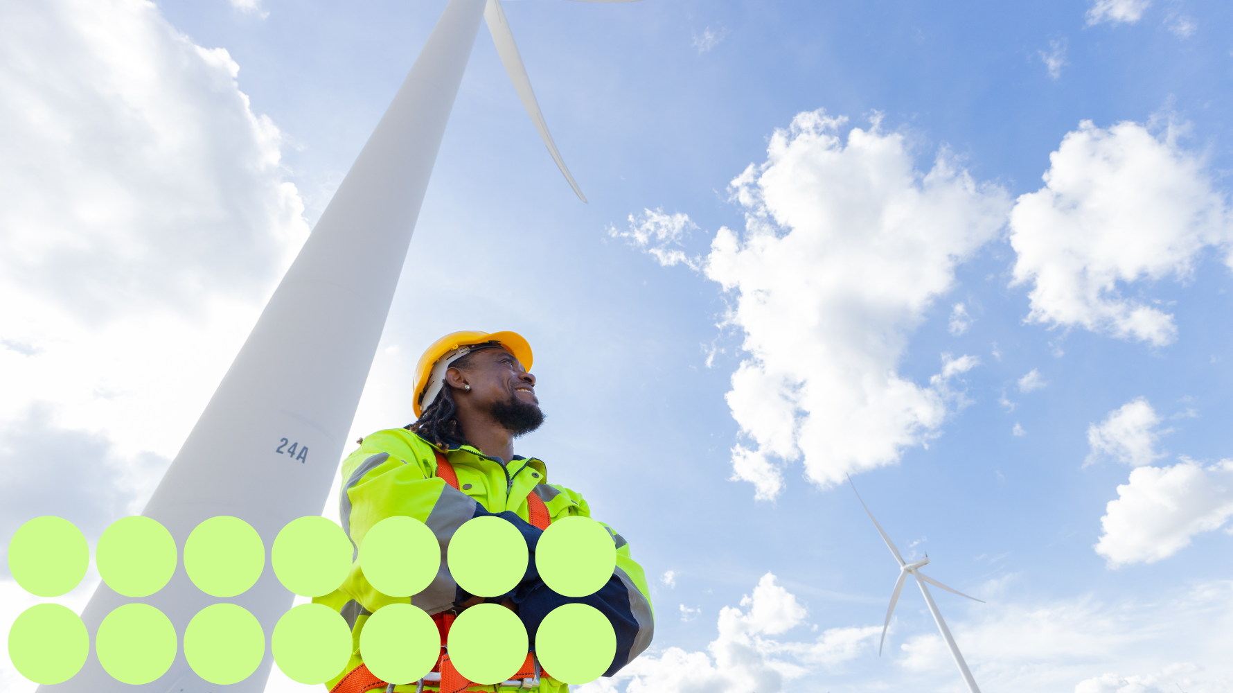 Man in safety vest and hard hat staring up at a blue sky with minimal clouds. Windmills are visible above him. 14 green circles are in the bottom left of the image for decoration.