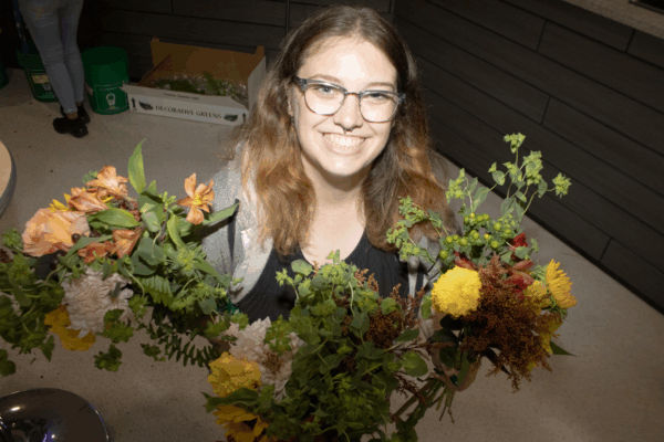 A smiling student holds two colourful floral arrangements with orange, yellow, and green blooms during a bouquet-making activity.