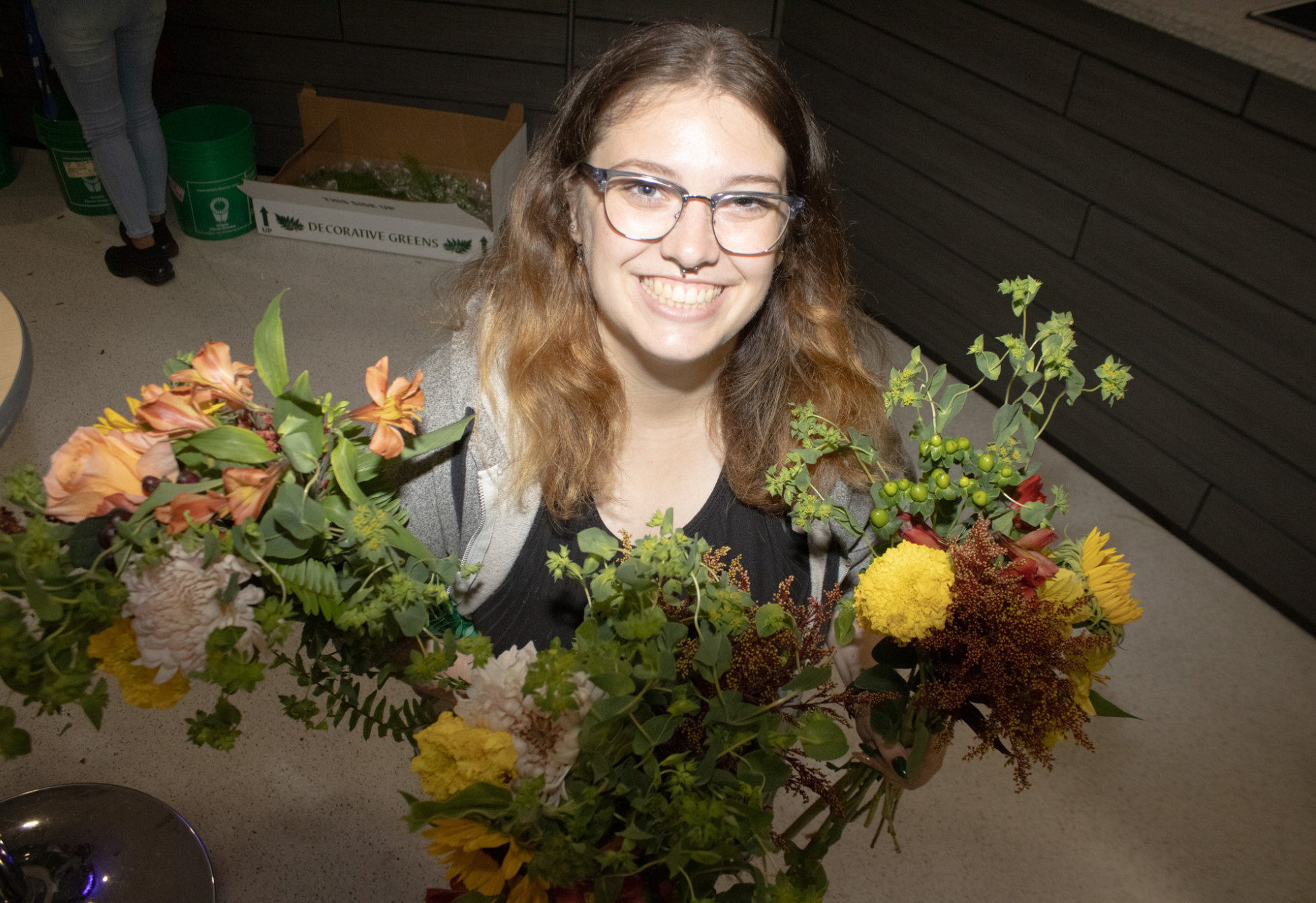 A smiling student holds two colourful floral arrangements with orange, yellow, and green blooms during a bouquet-making activity.