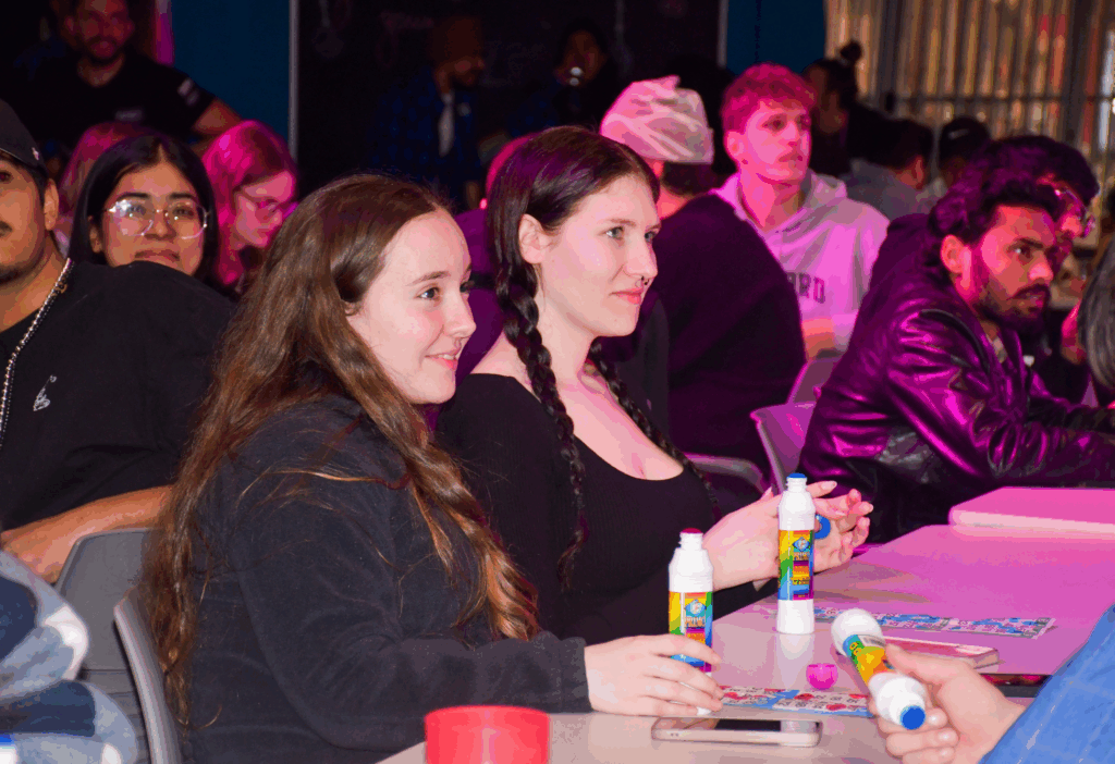 Two students sit at a table laughing and holding bingo dabbers during a lively drag bingo night.