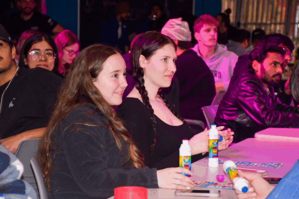 Two students sit at a table laughing and holding bingo dabbers during a lively drag bingo night.