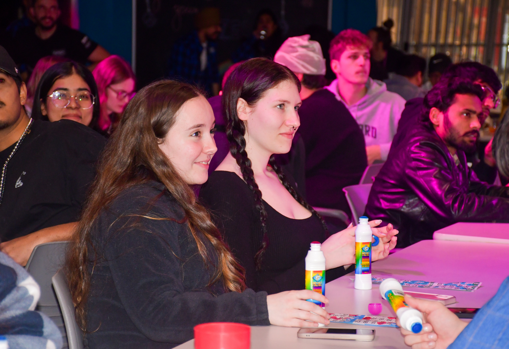 Two students sit at a table laughing and holding bingo dabbers during a lively drag bingo night.