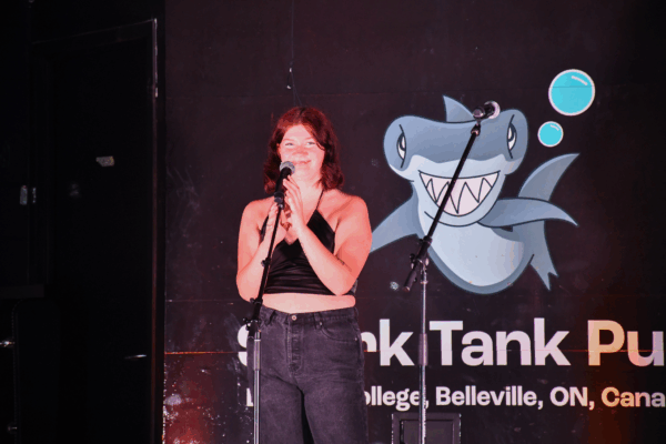 A student stands on stage with a microphone, smiling under the lights in front of a Shark Tank Pub backdrop.