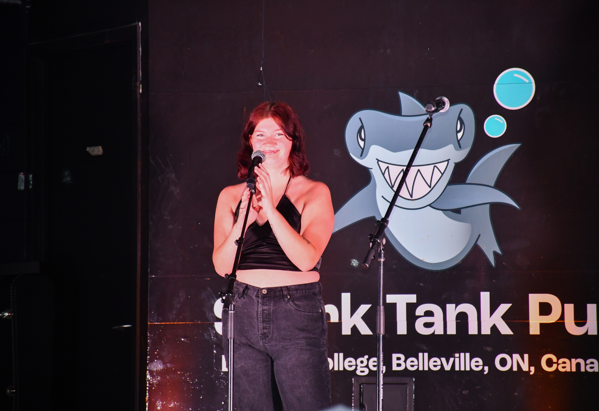 A student stands on stage with a microphone, smiling under the lights in front of a Shark Tank Pub backdrop.