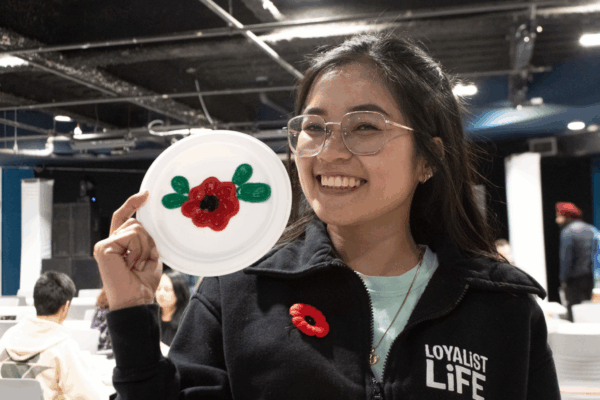 A student wearing a Loyalist Life jacket smiles while holding up a paper plate decorated with a red poppy design.