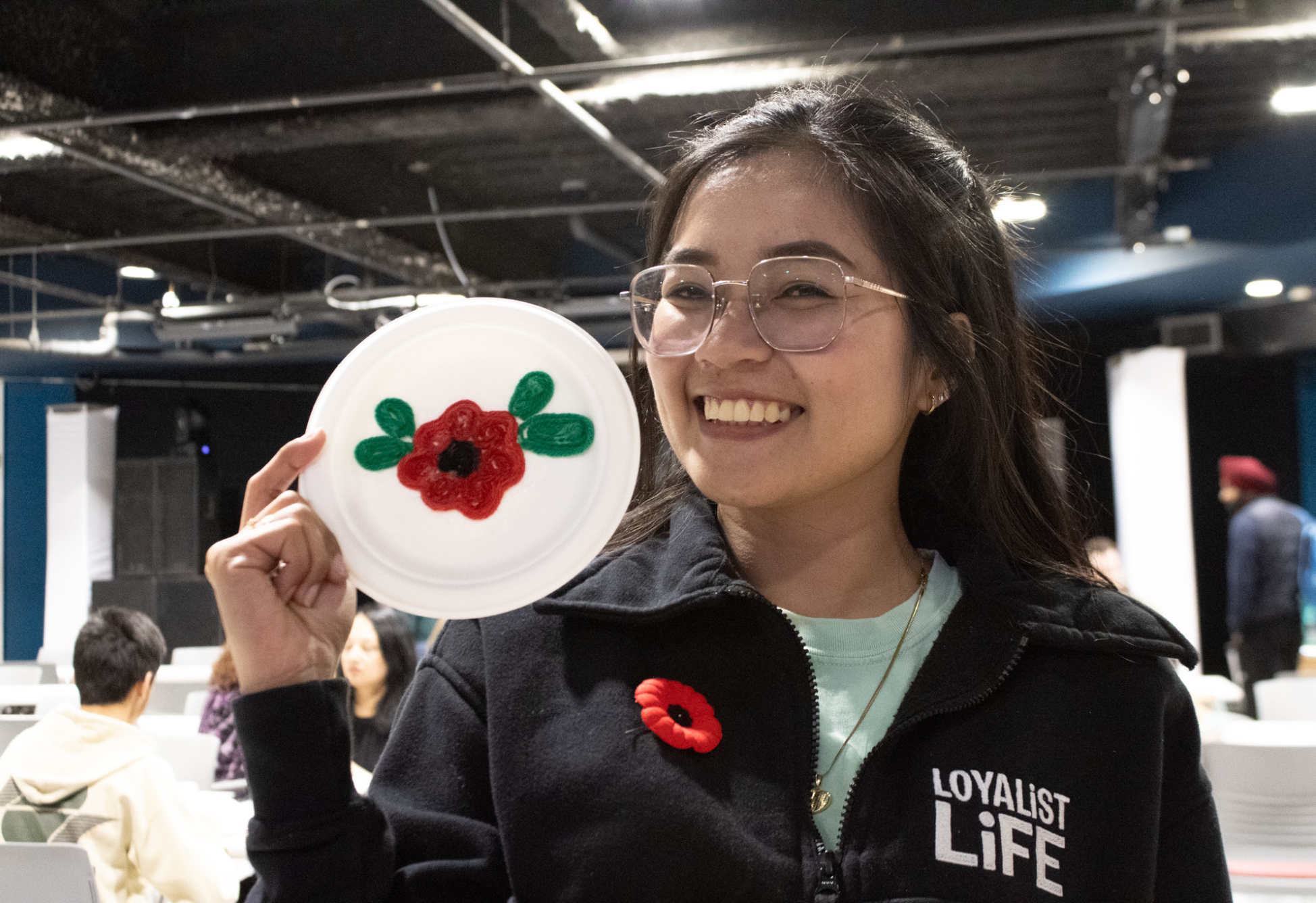 A student wearing a Loyalist Life jacket smiles while holding up a paper plate decorated with a red poppy design.